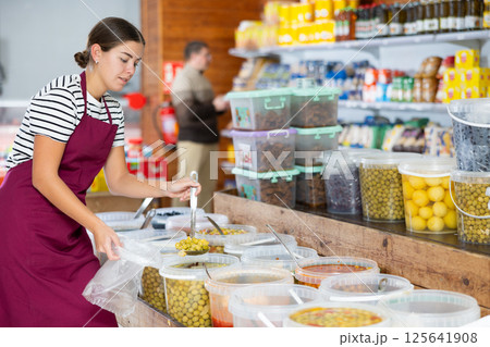 Young groceress scooping marinated olives into bag for customer at store Young groceress scooping marinated olives into bag for customer at store 125641908