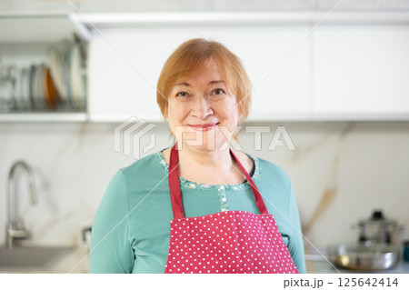 Elderly smiling lady in apron stands in clean kitchen and comes up with menu for lunch 125642414
