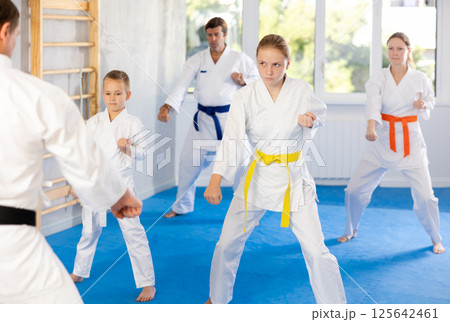 Teen girl dressed in white kimono with belt practice punches techniques with trainer, in background her family is doing kata 125642461