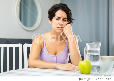 Dissatisfied young woman sitting at the table in room of her apartment Dissatisfied young woman sitting at the table in room of her apartment 125642637
