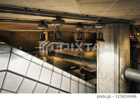 Industrial looking architecture, metal lamps shine over tubes, concrete walls and pillars in modern train station. Blurred people going down the escalator stairs 125650496