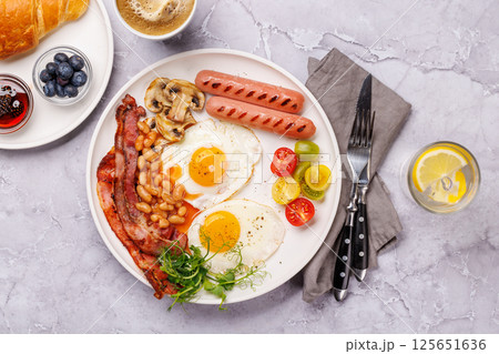 A classic breakfast plate featuring sunny-side-up eggs, crispy bacon, sausages, and fresh tomatoes 125651636