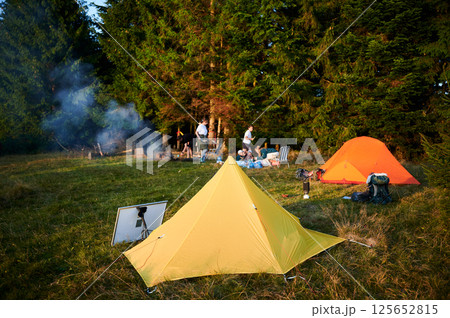Solar panel in grassy field with tents in background highlights eco-friendly camping setup. Scene includes campers, emphasizing integration of renewable energy in outdoor activities. Solar panel in grassy field with tents in background highlights eco-friendly camping setup. Scene includes campers, emphasizing integration of renewable energy in outdoor activities. 125652815