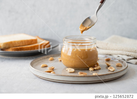Peanut butter flowing from spoon in glass jar on light background with salted nuts and bread toasts. Peanut butter flowing from spoon in glass jar on light background with salted nuts and bread toasts. 125652872