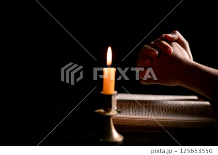 In the dark, hand clasped in prayer, illuminated by the soft candlelight, as black flames of candle fire flickered gently. candle, pray, hand, prayer, black, fire, flames, candlelight, light, dark. 125653550