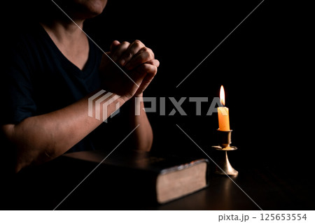 In the dark, hand clasped in prayer, illuminated by the soft candlelight, as black flames of candle fire flickered gently. candle, pray, hand, prayer, black, fire, flames, candlelight, light, dark. In the dark, hand clasped in prayer, illuminated by the soft candlelight, as black flames of candle fire flickered gently. candle, pray, hand, prayer, black, fire, flames, candlelight, light, dark. 125653554