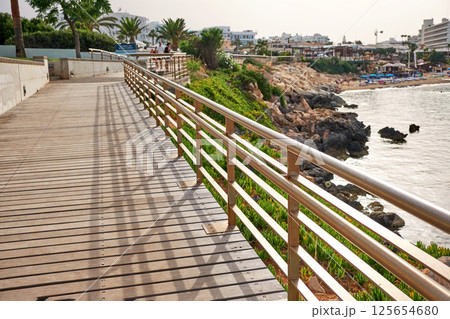 A wooden boardwalk with metal railing leads along the coast, offering a scenic view of the sea. A wooden boardwalk with metal railing leads along the coast, offering a scenic view of the sea. 125654680