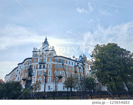A majestic historic building with a clock tower, white detailing, and ornate architecture rises against a blue sky, surrounded by lush trees and an urban park. The image highlights elegance and urban 125654883