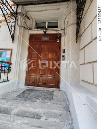 A sturdy wooden door with square panels, surrounded by an aged white facade, stone steps, and wrought iron canopy supports. The image highlights urban entrances and vintage architectural charm. A sturdy wooden door with square panels, surrounded by an aged white facade, stone steps, and wrought iron canopy supports. The image highlights urban entrances and vintage architectural charm. 125654884