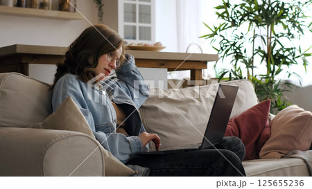 A focused woman in a denim jacket works from her couch, with a laptop and notes in front of her. This photograph promotes the idea of efficient and productive home-based workspaces. 125655236