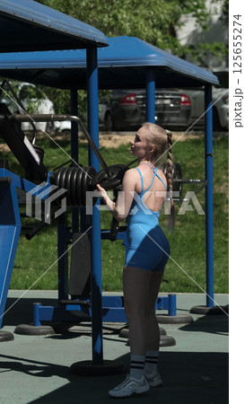 A woman in a blue bodysuit adjusts weights on exercise equipment at an outdoor gym, emphasizing her focus on building strength and endurance. This image connects the concept of outdoor workouts with 125655274