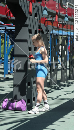 A woman in blue sportswear rehydrates with a water bottle during her outdoor workout session, standing by gym equipment. The photo showcases the crucial practice of drinking water to stay energized 125655275