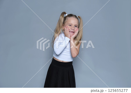 little girl with blonde pigtails stands with her hands on her cheeks, showing off a playful, surprised expression. This studio shot captures the whimsy and light-heartedness of childhood, paired with 125655277
