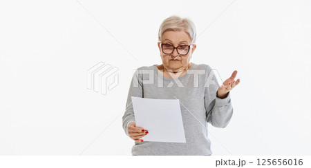Senior woman shrugging while holding sheet of paper against white studio background. Confused or uncertain reaction, online task Senior woman shrugging while holding sheet of paper against white studio background. Confused or uncertain reaction, online task 125656016