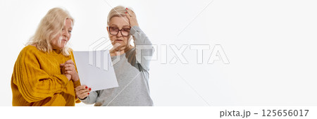 Banner. Two senior women looking at paper with worried expression against white studio background with negative space. Test results , real emotion concept. 125656017