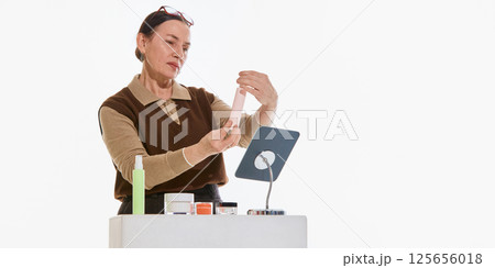 Mature woman checking skincare product near mirror against white background. Concept of beauty routine, 125656018