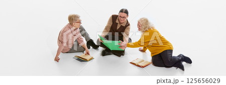 Banner, flyer. Three senior women reading and discussing books on floor against white studio background. Group study. Knowledge sharing 125656029