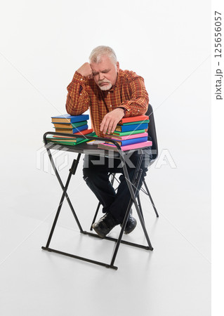 Elderly man resting on desk surrounded by books, showing exhaustion from studying, against white studio background. Elderly man resting on desk surrounded by books, showing exhaustion from studying, against white studio background. 125656057
