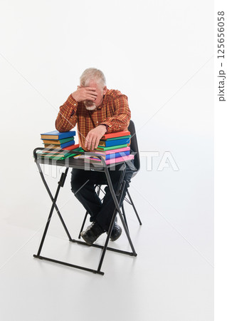 Older man overwhelmed by studies, hand on forehead, sitting with colorful books against white studio background. 125656058