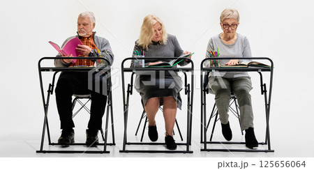 Three elderly students sitting at desks against white background, engaged with books and notes, showing active learning, teamwork, knowledge pursuit. 125656064