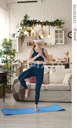 A woman in a teal outfit practices a balancing pose while wearing a VR headset in her stylish living room. This image highlights the innovative use of virtual reality technology in home fitness A woman in a teal outfit practices a balancing pose while wearing a VR headset in her stylish living room. This image highlights the innovative use of virtual reality technology in home fitness 125656706