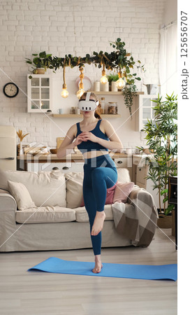 Engaged in a virtual reality workout, a woman in teal activewear balances on one foot in her modern living room. This photo captures the dynamic integration of VR technology with exercise. Engaged in a virtual reality workout, a woman in teal activewear balances on one foot in her modern living room. This photo captures the dynamic integration of VR technology with exercise. 125656707
