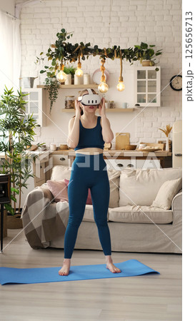 Balancing on one foot, a woman in teal workout clothes engages in a VR fitness session in her stylish living room. The photograph promotes the innovative blend of technology and exercise in Balancing on one foot, a woman in teal workout clothes engages in a VR fitness session in her stylish living room. The photograph promotes the innovative blend of technology and exercise in 125656713