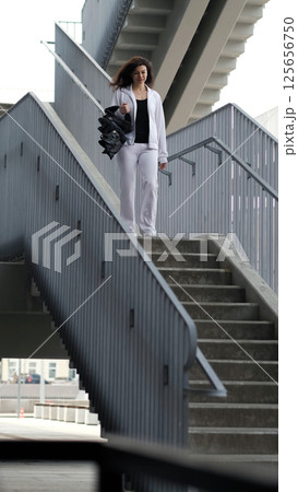 A woman dressed in sportswear moves down a metal staircase, carrying specialized jumping boots. The industrial backdrop emphasizes the fusion of fitness, architecture, and city movement. 125656750