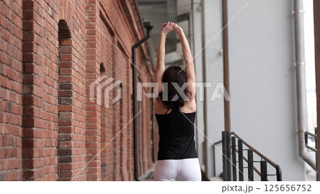 A woman in sportswear stretches her arms upward while standing near a brick wall. The blend of fitness and urban architecture symbolizes balance, movement, and well-being in modern city life. A woman in sportswear stretches her arms upward while standing near a brick wall. The blend of fitness and urban architecture symbolizes balance, movement, and well-being in modern city life. 125656752