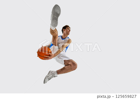Low angle view. Athletic young man in white-blue game outfit sprinting with basketball, practicing dribbling technique with intensity against white background. 125659827