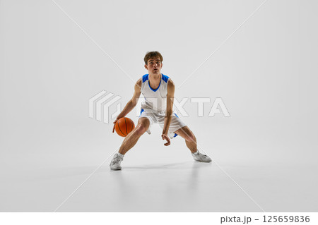 ale athlete wearing white-blue uniform in wide-leg stance, dribbling ball with determination and ready to break through defense against white background. ale athlete wearing white-blue uniform in wide-leg stance, dribbling ball with determination and ready to break through defense against white background. 125659836