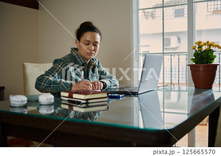 Focused Woman Writing Notes at a Desk with Laptop in Home Office 125665570