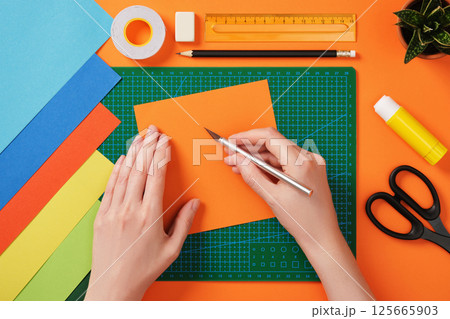 Hands working with colorful paper using a precision knife on a green cutting mat. Tools for paper crafts. Ruler, pencil, glue stick, scissors, tape, eraser 125665903