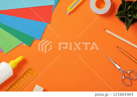 Supplies for paper art hobby: scissors, craft knife, pencil, eraser, colored paper on orange background. Space for your text. Paper crafts work table flatlay. 125665905