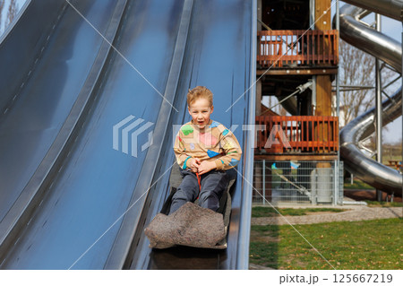 Cute adorable little kid blond boy enjoy riding fast on playground Highrise metal slide with fabric mat at city nature park recreation area on sunny day. Outside children summer spring activity Cute adorable little kid blond boy enjoy riding fast on playground Highrise metal slide with fabric mat at city nature park recreation area on sunny day. Outside children summer spring activity 125667219