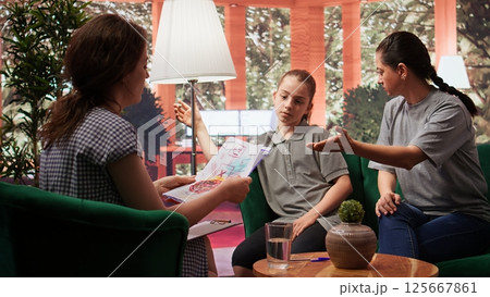Therapist presenting unsettling dangerous drawings to mother, teen girl suffering from depersonalization and derealization. Teenager with dissociation and loss of contact at therapy. Camera B. Therapist presenting unsettling dangerous drawings to mother, teen girl suffering from depersonalization and derealization. Teenager with dissociation and loss of contact at therapy. Camera B. 125667861