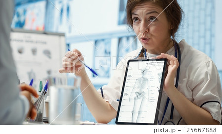 Healthcare provider presenting lab test results to a patient in cabinet, reviewing x ray and MRI scans to assist man through recovery. Medical technology in diagnostics and care management. Camera A. 125667888