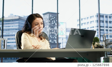 Young entrepreneur listening to a podcast while she solves daily business tasks in high end office, corporate success story. Female director working with headphones, determination. Camera A. Young entrepreneur listening to a podcast while she solves daily business tasks in high end office, corporate success story. Female director working with headphones, determination. Camera A. 125667898