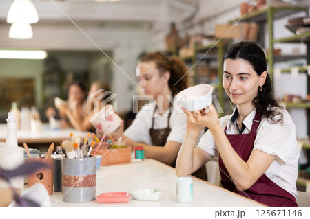 Young woman sits near table and decorates clay ceramic craft vase, plate Young woman sits near table and decorates clay ceramic craft vase, plate 125671146