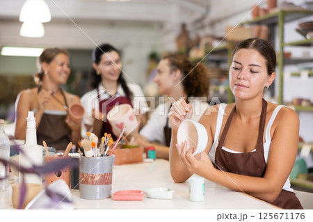 Young woman sits near table and decorates clay ceramic craft vase, plate Young woman sits near table and decorates clay ceramic craft vase, plate 125671176