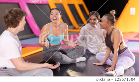 Group of cheerful sporty young people sharing impressions while sitting on trampoline in modern leisure sports center Group of cheerful sporty young people sharing impressions while sitting on trampoline in modern leisure sports center 125671304