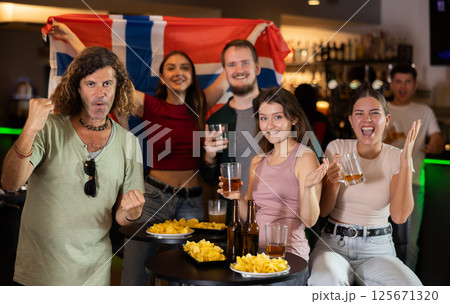 Group of friends, excited supporters cheering for favorite team with flag of Norway while watching match on TV 125671320