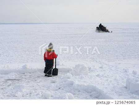Little boy with a shovel in a huge space covered with snow and ice watches a snowmobile whizzing by 125674065