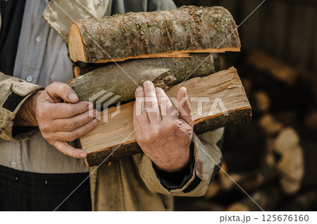 Rough, veined hands carry freshly chopped logs. Behind them, firewood is piled high. 125676160