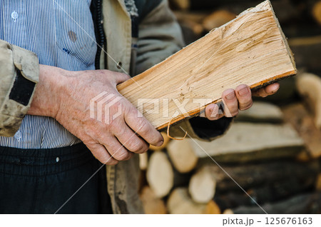 Calloused hands hold split wood tightly. In the background, stacked firewood is neatly arranged. Calloused hands hold split wood tightly. In the background, stacked firewood is neatly arranged. 125676163
