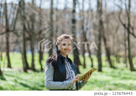 A beautiful girl in leather pants and bomber jacket with a tablet in a spring park. Happy woman with a tablet in her hands smiling sweetly. A beautiful girl in leather pants and bomber jacket with a tablet in a spring park. Happy woman with a tablet in her hands smiling sweetly. 125676443