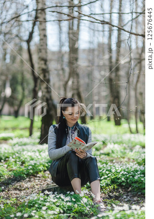 A girl in a spring park resting with a book in her hands. A young woman is sitting on the grass in a green forest.  125676445