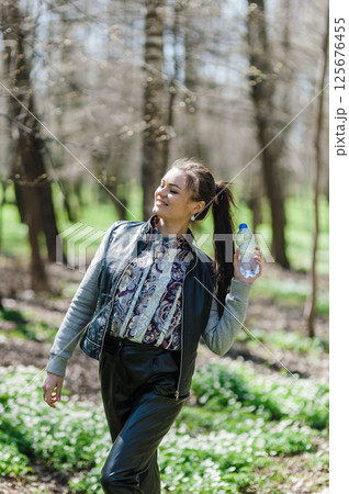 A beautiful girl in leather pants and a bomber jacket holds a bottle of water in a spring forest and basks in the sun. A young woman is walking in the park with a bottle of mineral water. 125676455