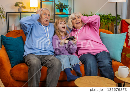 Child girl granddaughter grandparents using joystick controller playing console videogame at home 125676878