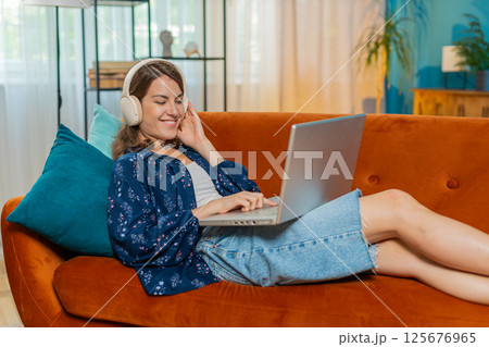 Happy woman in wireless headphones lying on home couch listening music working on laptop relaxing Happy woman in wireless headphones lying on home couch listening music working on laptop relaxing 125676965
